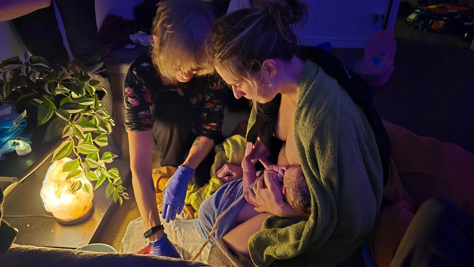 Ett nyfött barn vilar i mammans famn medan en barnmorska hjälper till, i varmt lampljus hemma.