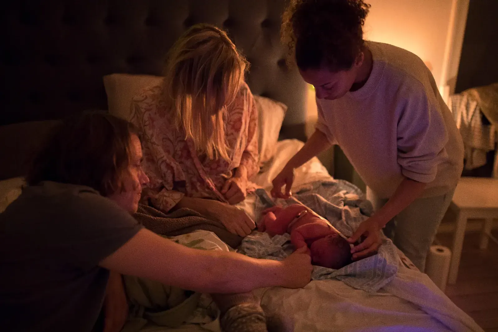 Hembarnmorskan undersöker den nyfödda bebisen i sängen i varmt lampljus medan föräldrarna ser på.