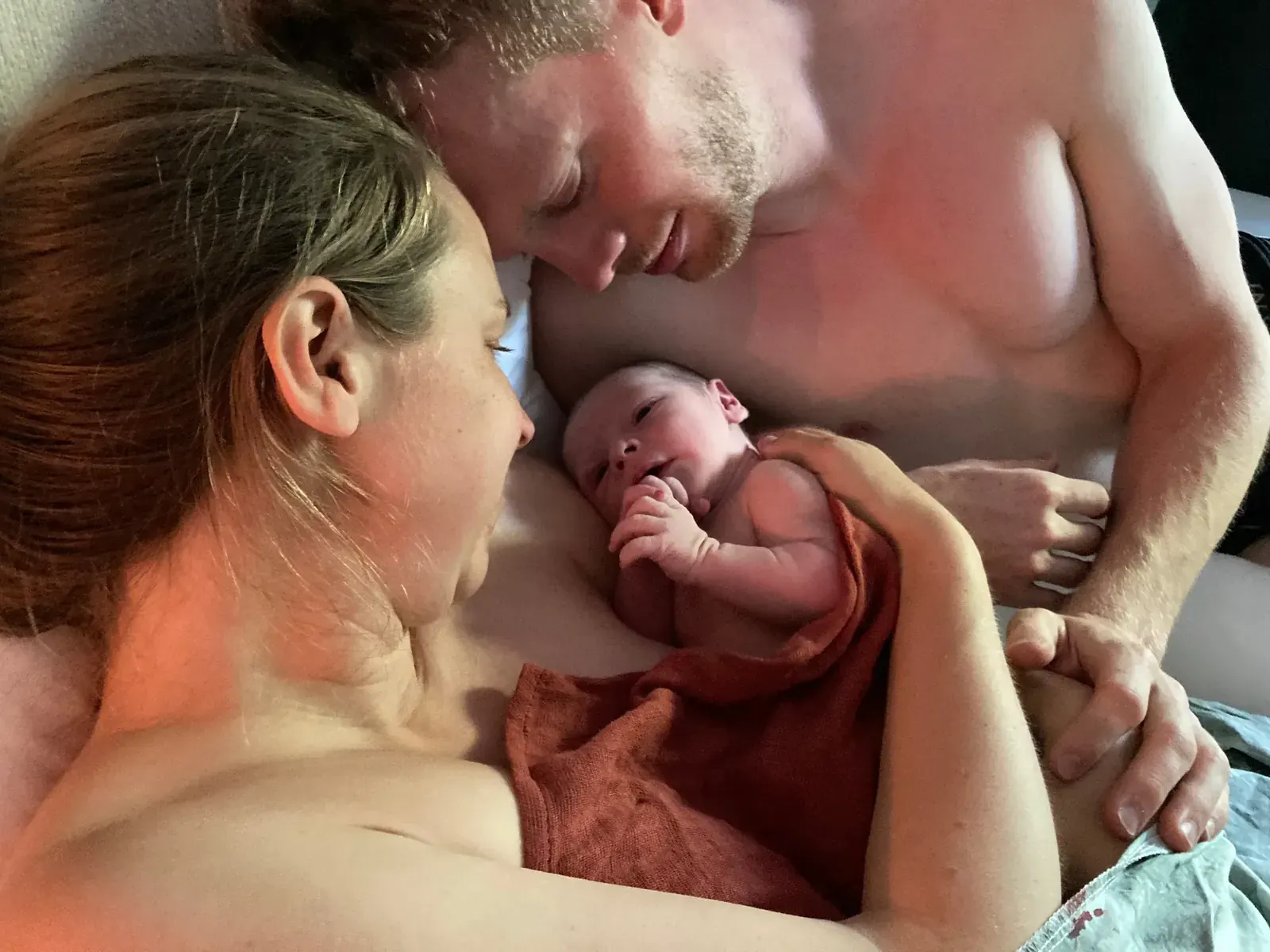 Nyfödd bebis insvept i orange handduk vilar mellan mamma och pappa hud mot hud i sängen direkt efter hemfödseln.