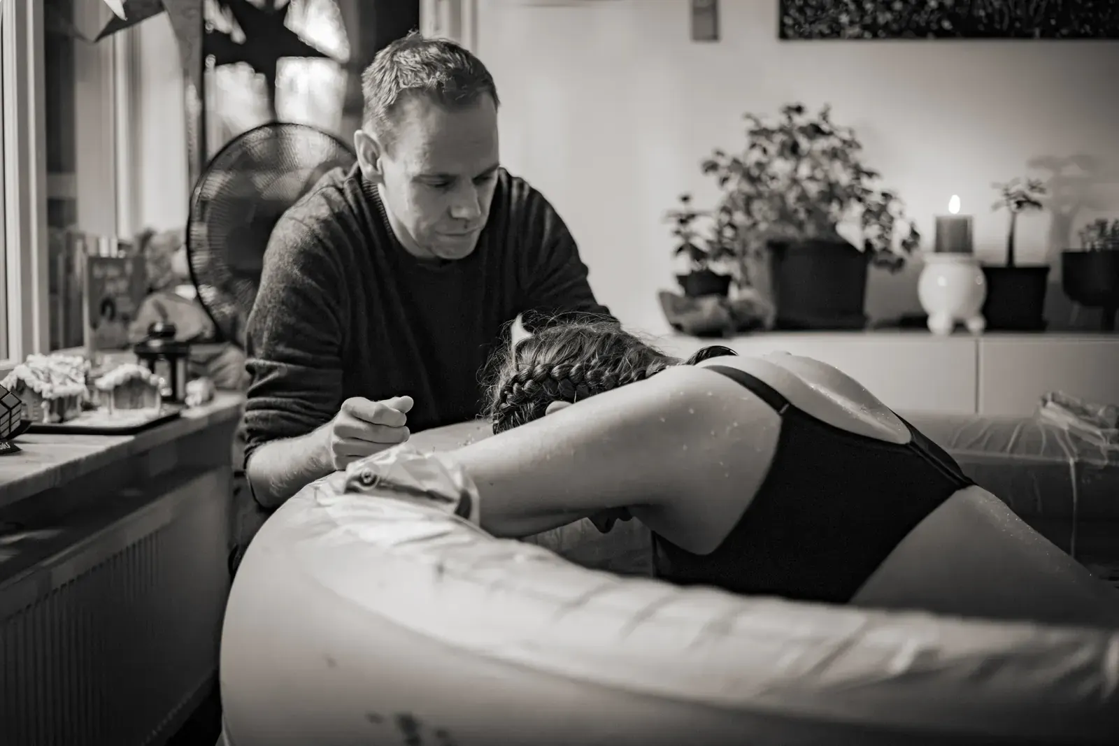 Svartvit bild av en kvinna som lutar sig framåt över kanten på en förlossningspool hemma medan partnern sitter bredvid och håller hennes hand under en värk.