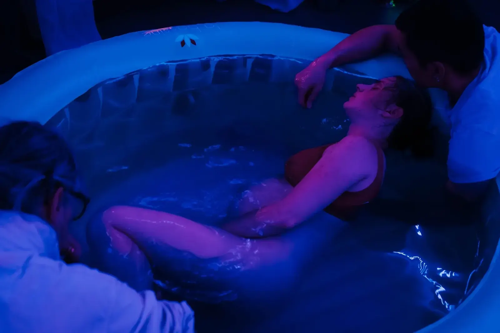 Jessica vilar tillbakalutad i förlossningspoolen i blått ljus medan Teddy håller om henne från poolkanten.