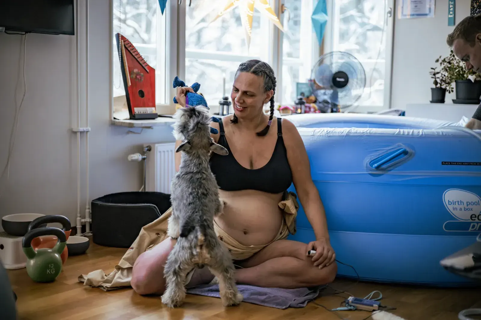 Gravid kvinna sitter på golvet bredvid en uppblåsbar förlossningspool och leker med sin hund.