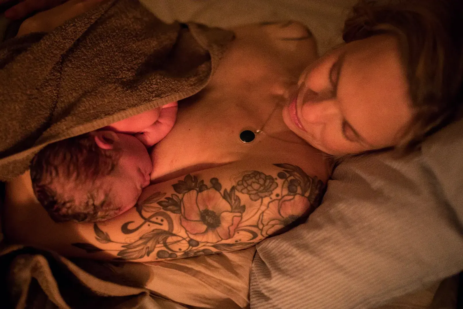 Nyfödd bebis vilar hud mot hud på mammans bröst, insvept i en handduk i varmt ljus.