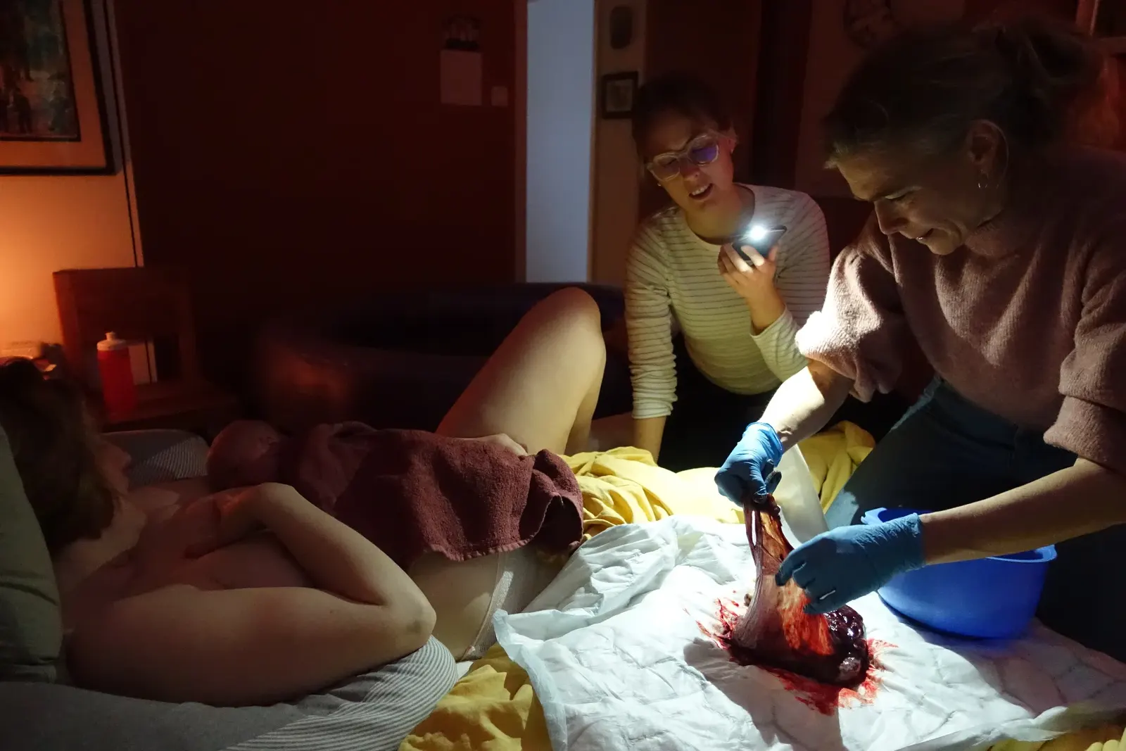 Hembarnmorskorna undersöker moderkakan i lampskenet medan Vera vilar bredvid med bebisen.