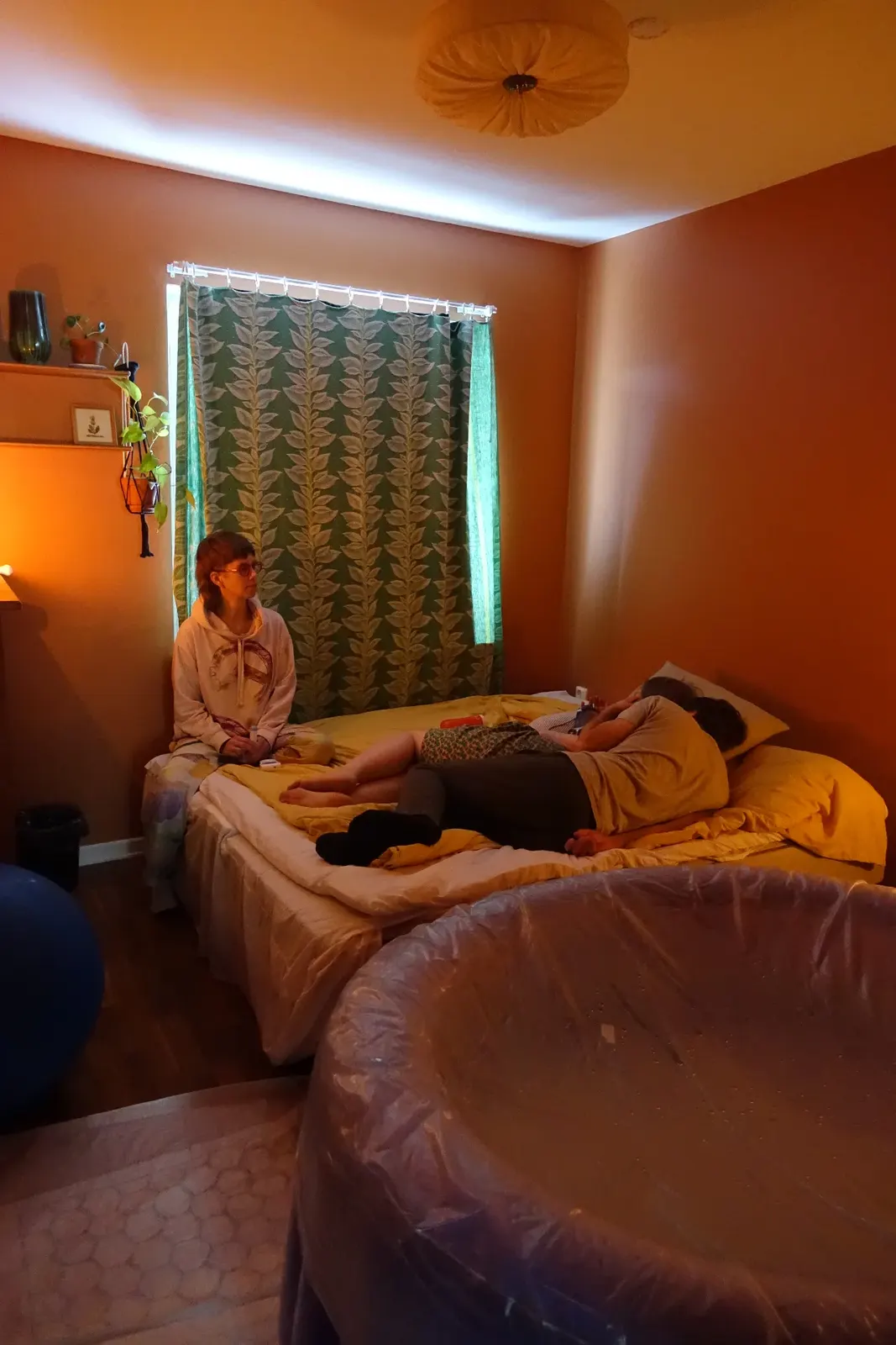 Sovrummet i varmt ljus med poolen klar i förgrunden, Vera vilar på sängen med sin partner medan barnmorskan sitter avvaktande bredvid.