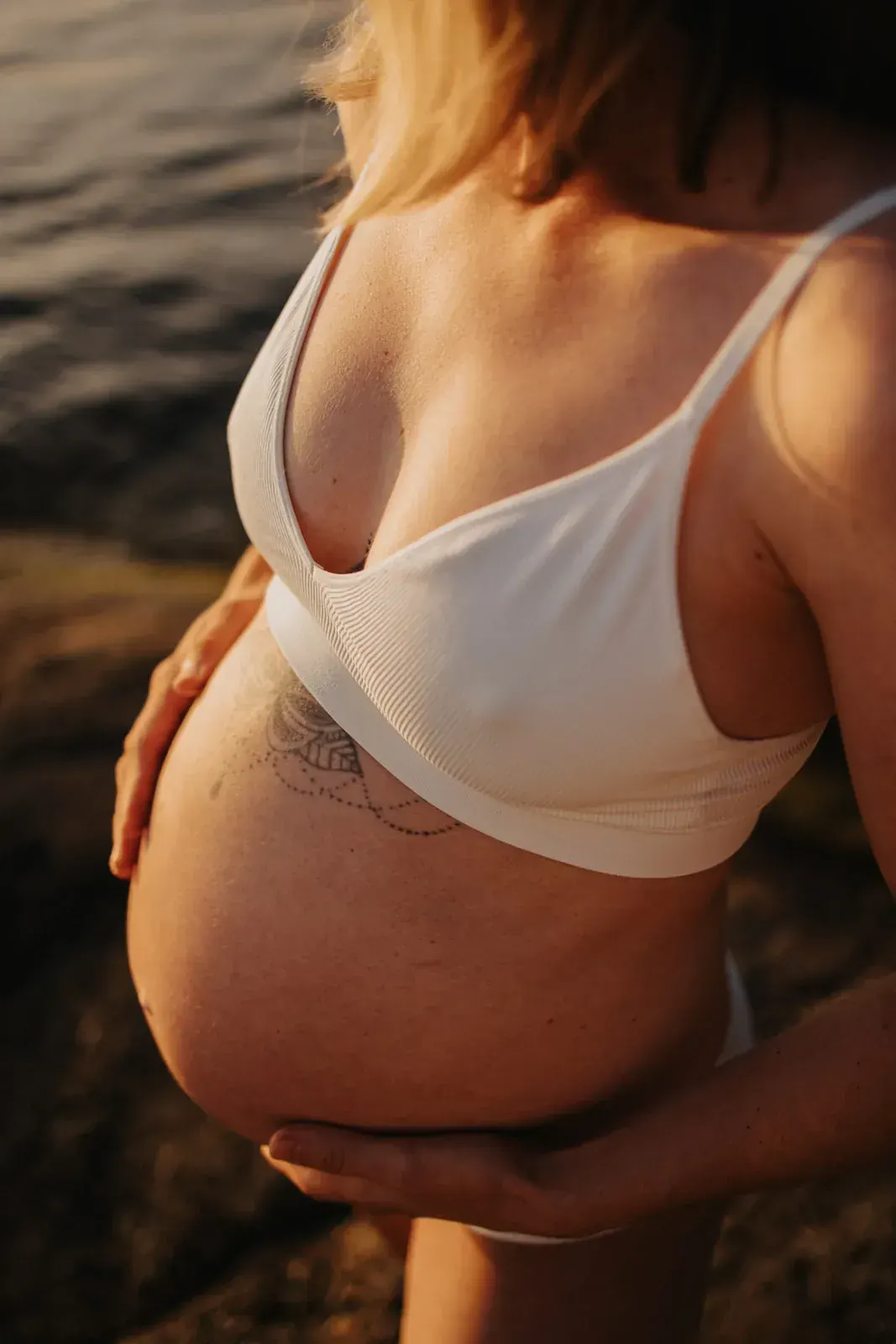 Närbild på en gravid kvinnas mage i varmt kvällsljus vid vatten.