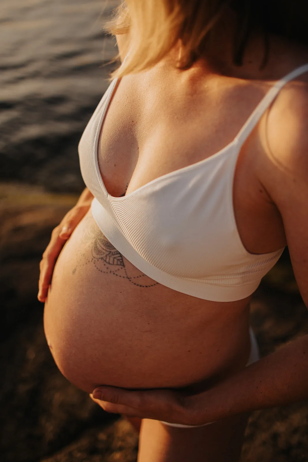 Närbild på en gravid kvinnas mage i varmt kvällsljus vid vatten.